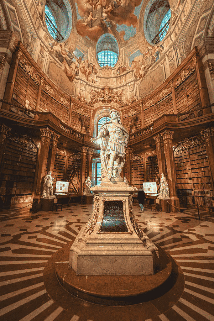 An ornate historic library has tall wooden bookshelves, a large statue in the center, and colorful ceiling frescoes above.