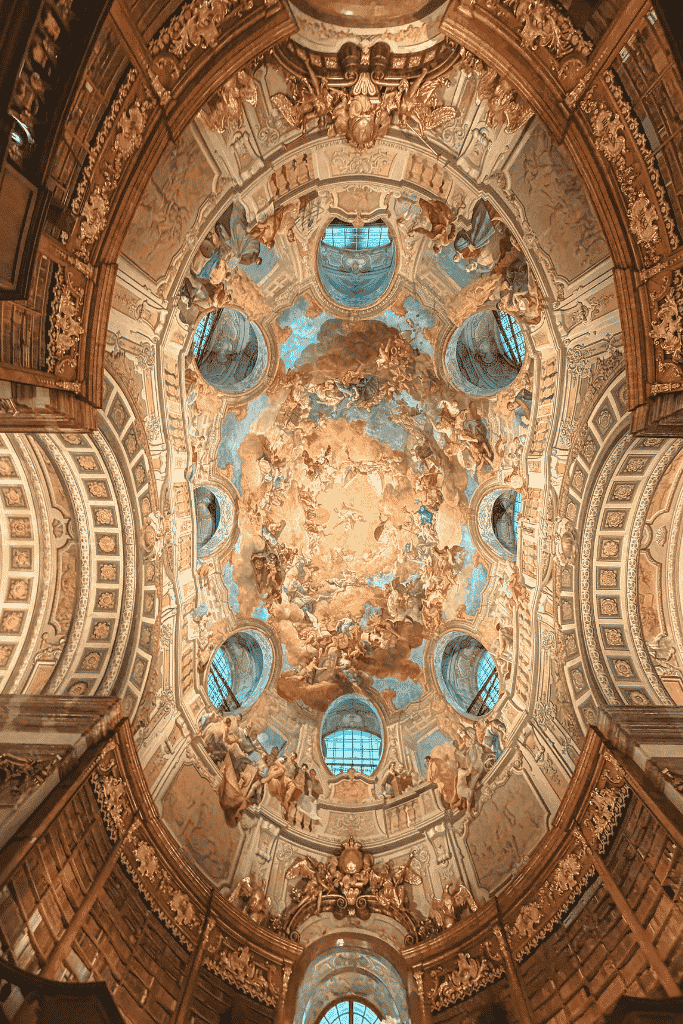 An ornate library ceiling is covered with detailed frescoes, gold decorations, and oval windows letting in natural light.