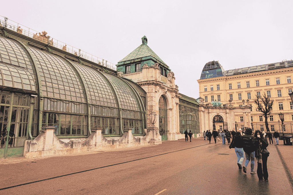 A large historic glass greenhouse with green metal framing stands beside an ornate palace as visitors walk along the path.