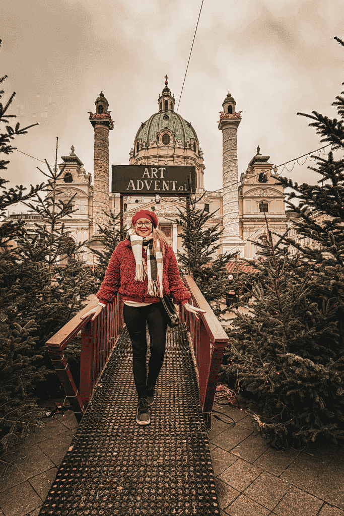 Kate in a red hat and jacket stands among Christmas trees at the Art Advent market with a large domed church behind her. A plate of crispy schnitzel is served at a table inside a bright glass greenhouse café filled with tropical plants and diners. A woman in a red hat and jacket stands among Christmas trees at the Art Advent market with a large domed church behind her.