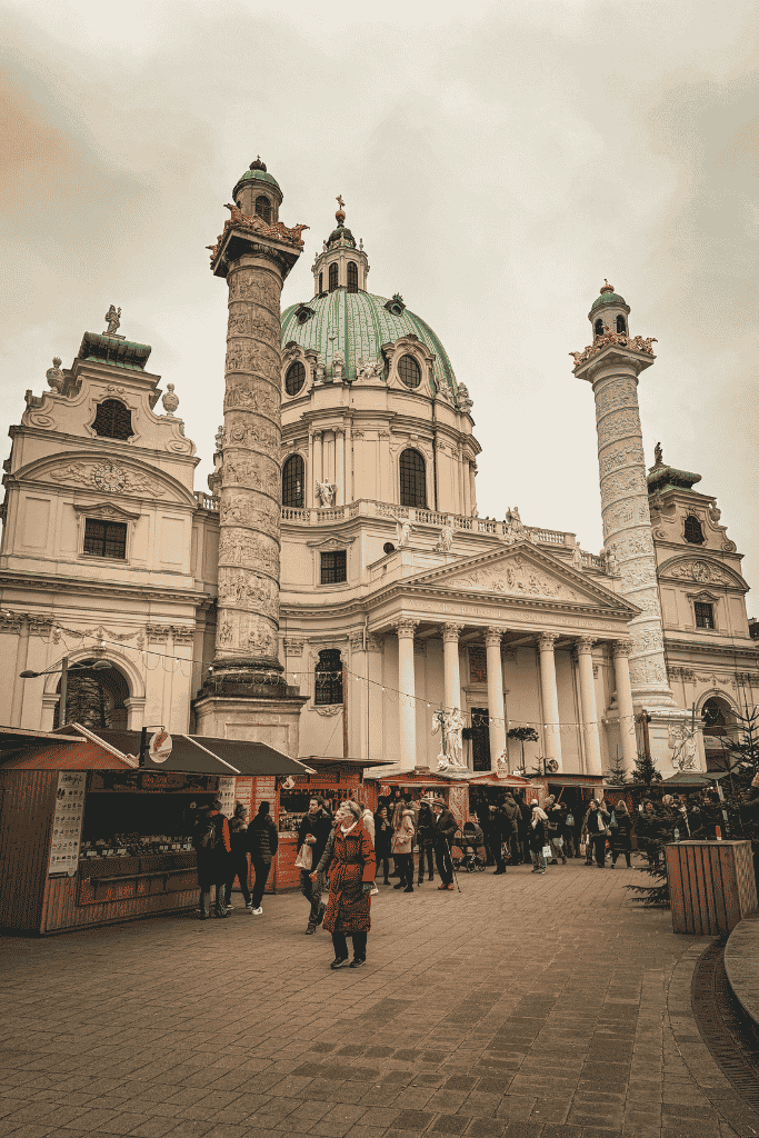 A Christmas market with wooden stalls sits in front of a grand domed church with two tall decorated columns.