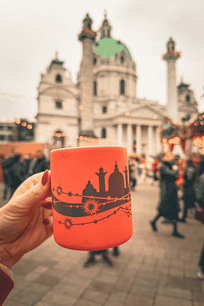 A hand holds a glowing orange Christmas market mug with a church design, with a grand domed church and festive lights in the background.