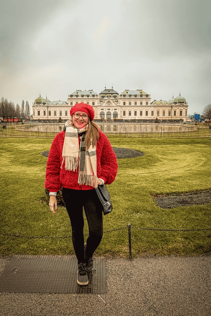 Kate in a red jacket and red hat stands in a garden in front of the grand Belvedere Palace in Vienna.