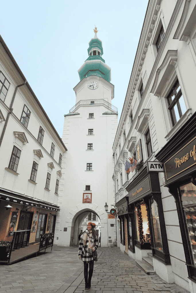 Kate stands on a cobblestone street beneath the white clock tower of Michael’s Gate in Bratislava.