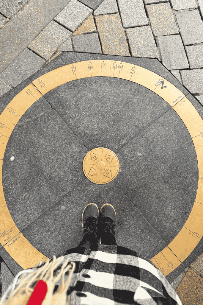 A person stands on a circular brass marker in the pavement showing distances and directions to cities around the world.