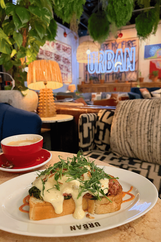 Eggs Benedict on toast topped with greens sits beside a red cup of coffee in a cozy, colorful hotel lounge.