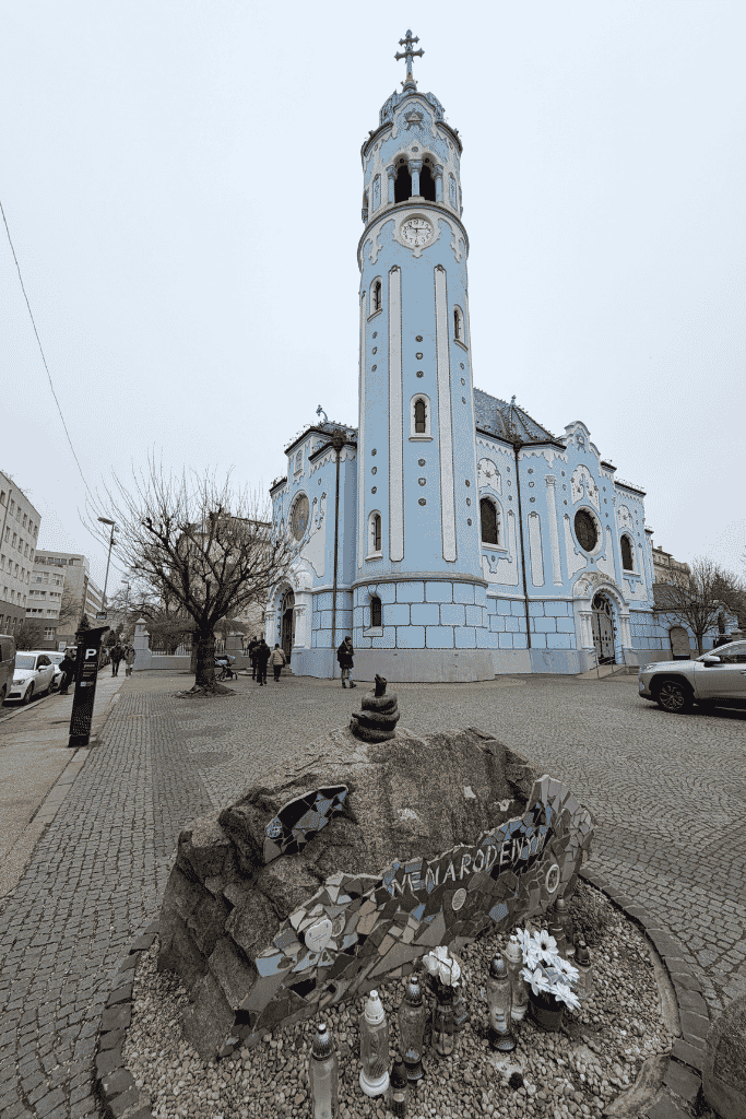 A pale blue church with a tall clock tower stands behind a small memorial covered with candles and flowers in Bratislava.