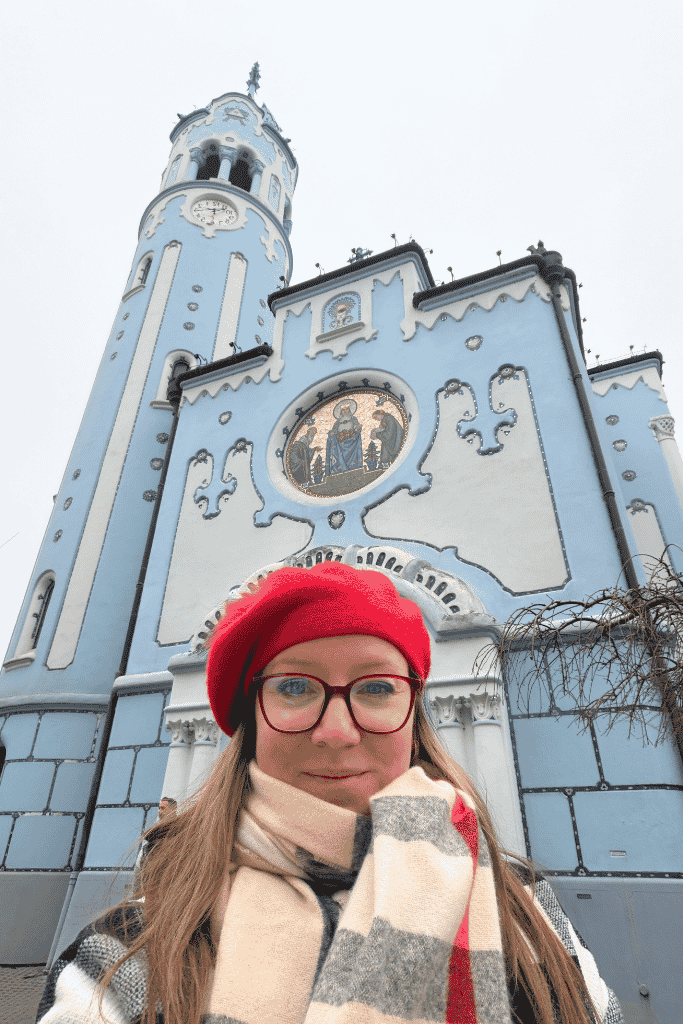 Kate in a red hat and warm scarf takes a selfie in front of the pale blue Church of St. Elizabeth in Bratislava.