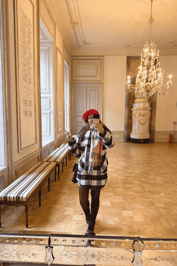 A woman in a red hat takes a mirror selfie inside an elegant palace room with a large crystal chandelier.
