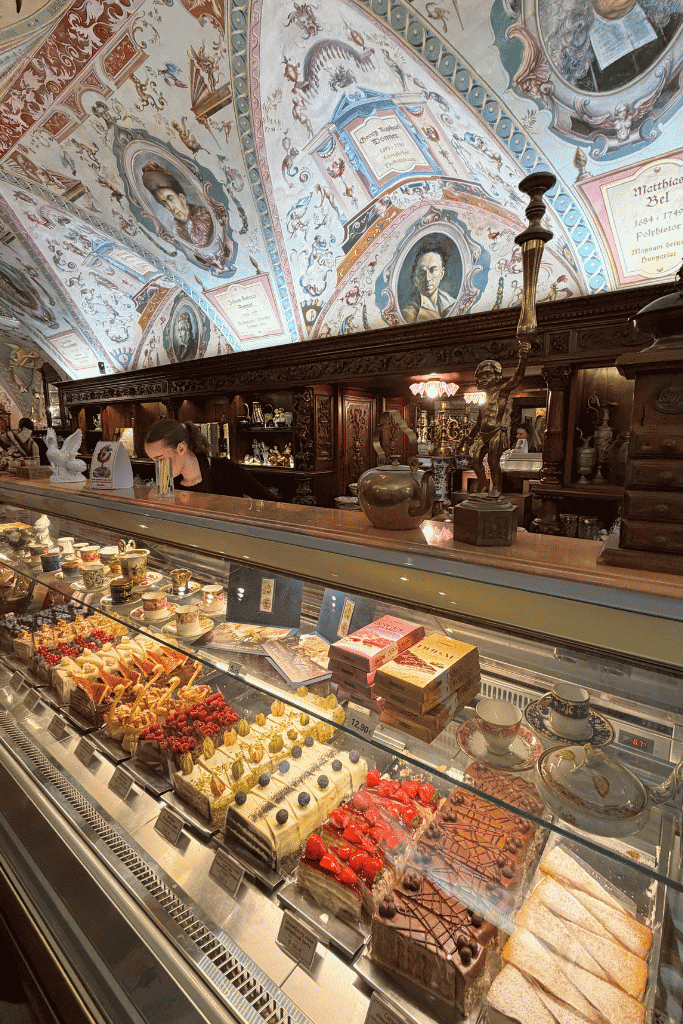 Ornate European café interior with a glass display case filled with colorful cakes and pastries beneath a frescoed ceiling.