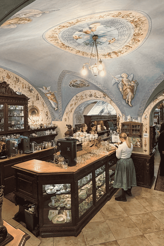 Alt text: Historic European café with an ornate frescoed ceiling, dark wood counters, glass pastry displays, and a barista behind the counter.