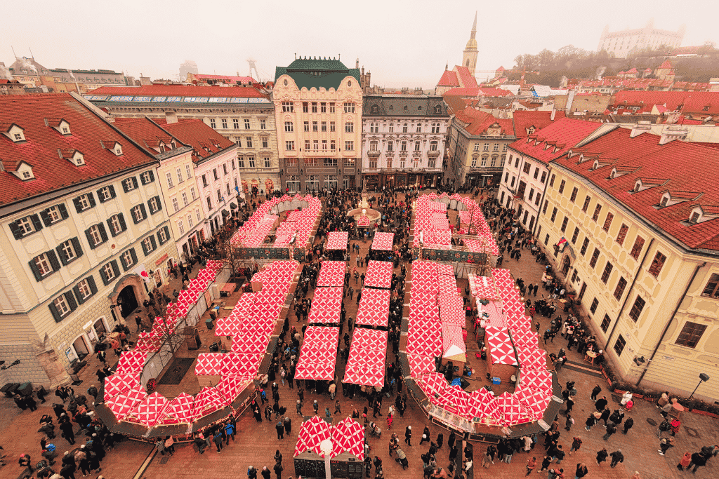 A view from above shows a crowded Christmas market with rows of red-and-white-roofed stalls filling Bratislava’s Main Square.