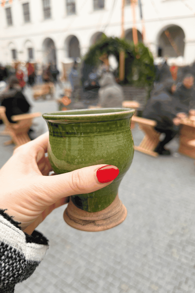 A hand with red nails holds a green ceramic mug at an outdoor Christmas market in Bratislava.