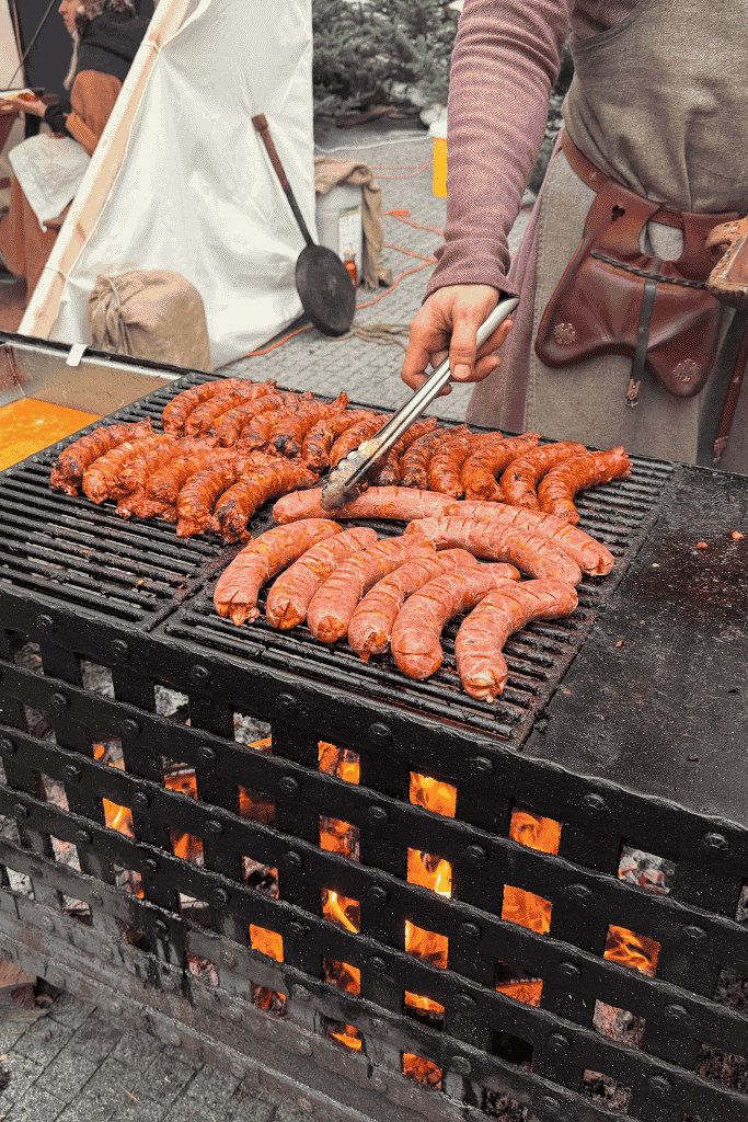 A vendor uses tongs to grill rows of sausages over an open fire at a Christmas market.