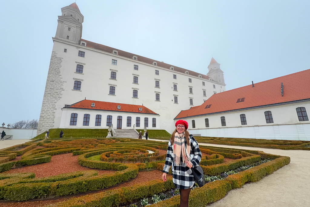 A woman in a red hat stands beside the formal gardens in front of Bratislava Castle.