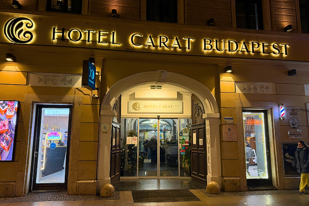 The warmly lit entrance of Hotel Carat Budapest at night, with the hotel name glowing above the doors.