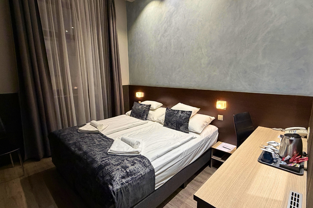 A simple Hotel Carat Budapest room with a double bed, bedside lights, dark curtains, and a desk with a kettle.