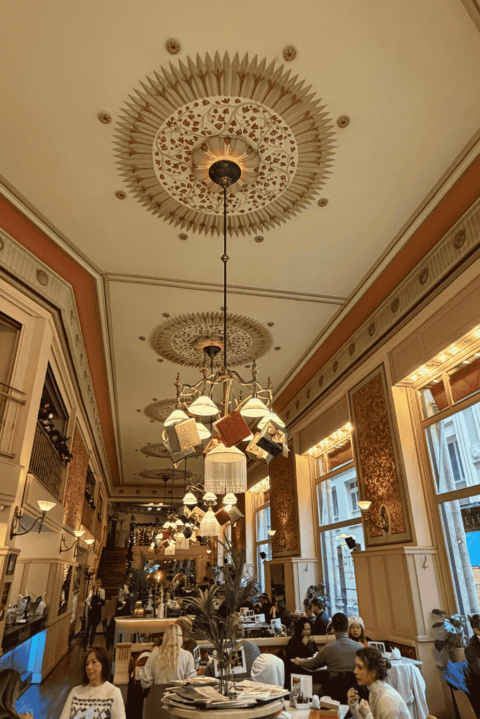 A busy, elegant café with high decorated ceilings, large windows, hanging lamps, and people seated at tables.
