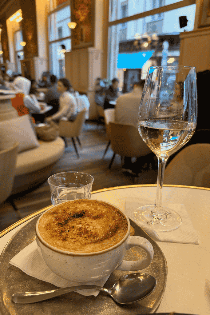 A cup of frothy coffee and a glass of water sit on a table inside a busy, elegant café.