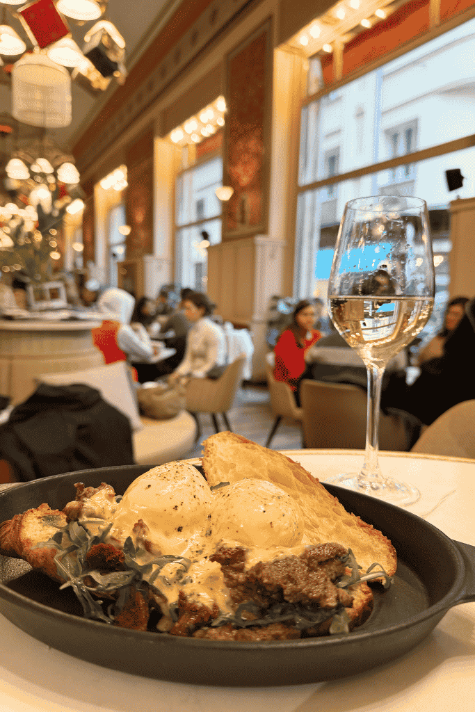 A skillet with eggs, toast, meat, and greens sits on a table inside a busy, elegant café.