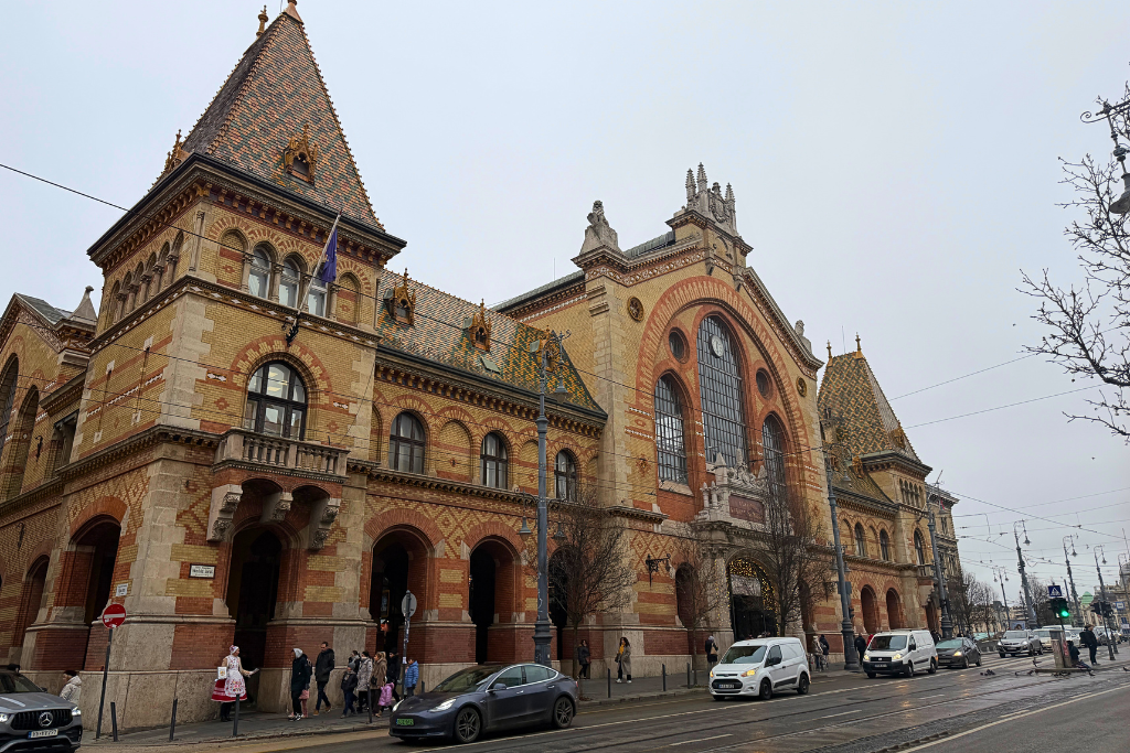 The historic Great Market Hall in Budapest stands beside a busy street, with its colorful tiled roof, tall windows, and brick facade.