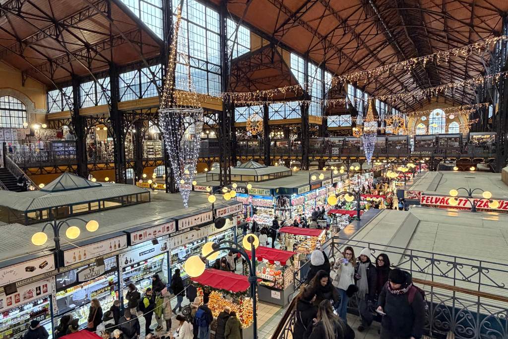 Inside Budapest’s Great Market Hall, shoppers walk among busy market stalls decorated with glowing Christmas lights.