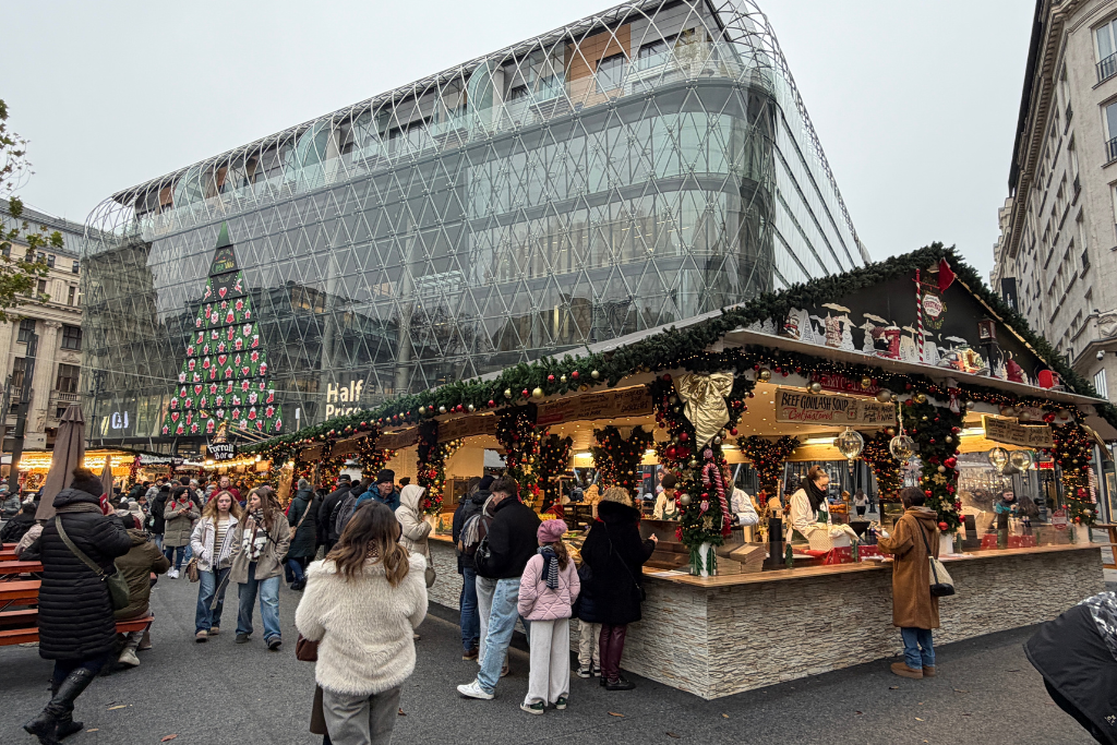 A busy Budapest Christmas market with decorated food stalls and crowds in front of a large modern glass building.