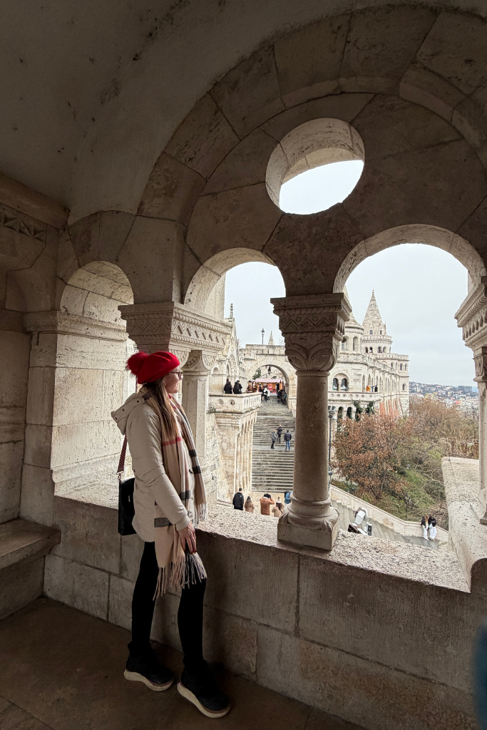 Kate in a red hat stands beneath a stone arch at Fisherman’s Bastion, looking out over the towers and stairways.