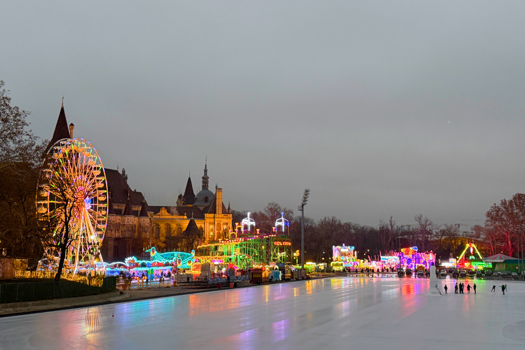 A colorful Ferris wheel and amusement rides glow beside a large outdoor ice-skating rink in Budapest at night.