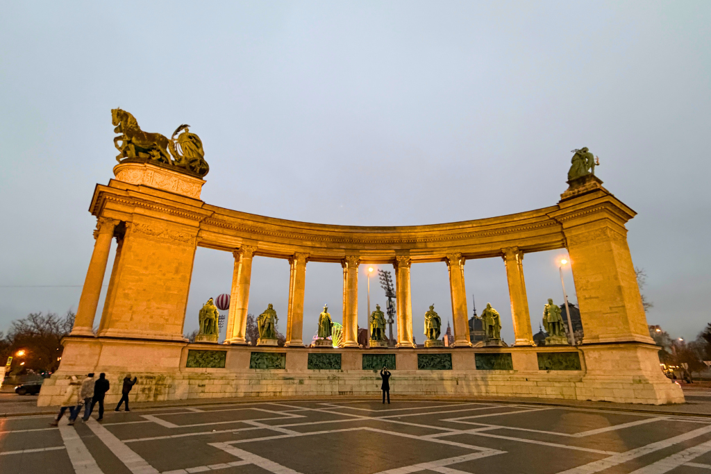 A curved stone monument with columns and statues glows at Heroes’ Square in Budapest at dusk.