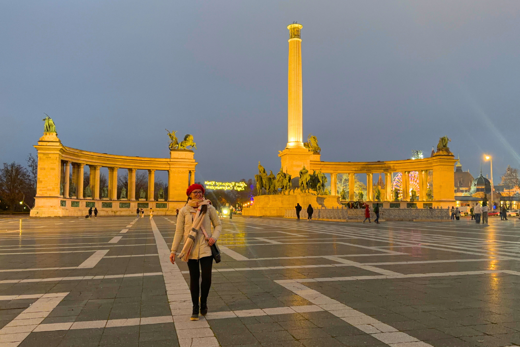 Kate in a red hat stands in front of the illuminated statues and tall central column at Heroes’ Square in Budapest.