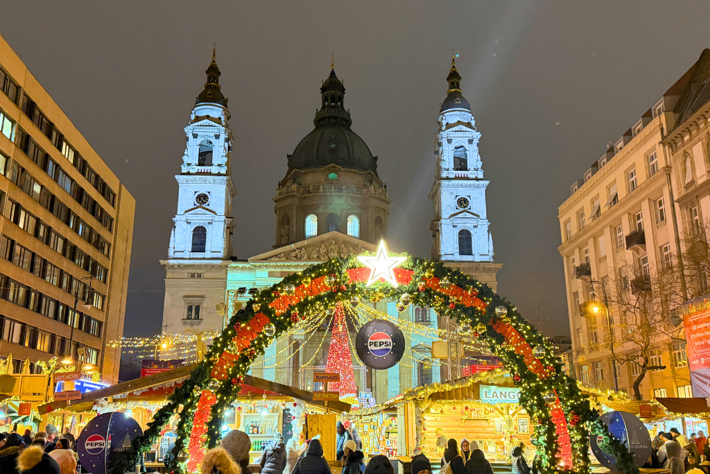 A busy Christmas market glows beneath a decorated arch in front of St. Stephen’s Basilica in Budapest at night.