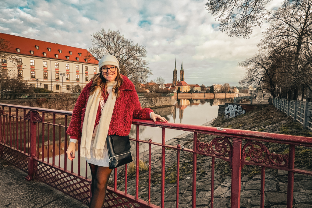 Kate is standing on an ornate red bridge beside a river, with historic architecture and twin church spires in the background.