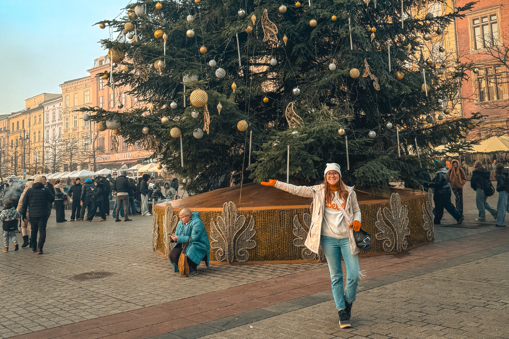 Kate posing beside a towering decorated Christmas tree in a lively European city square, surrounded by festive market activity.