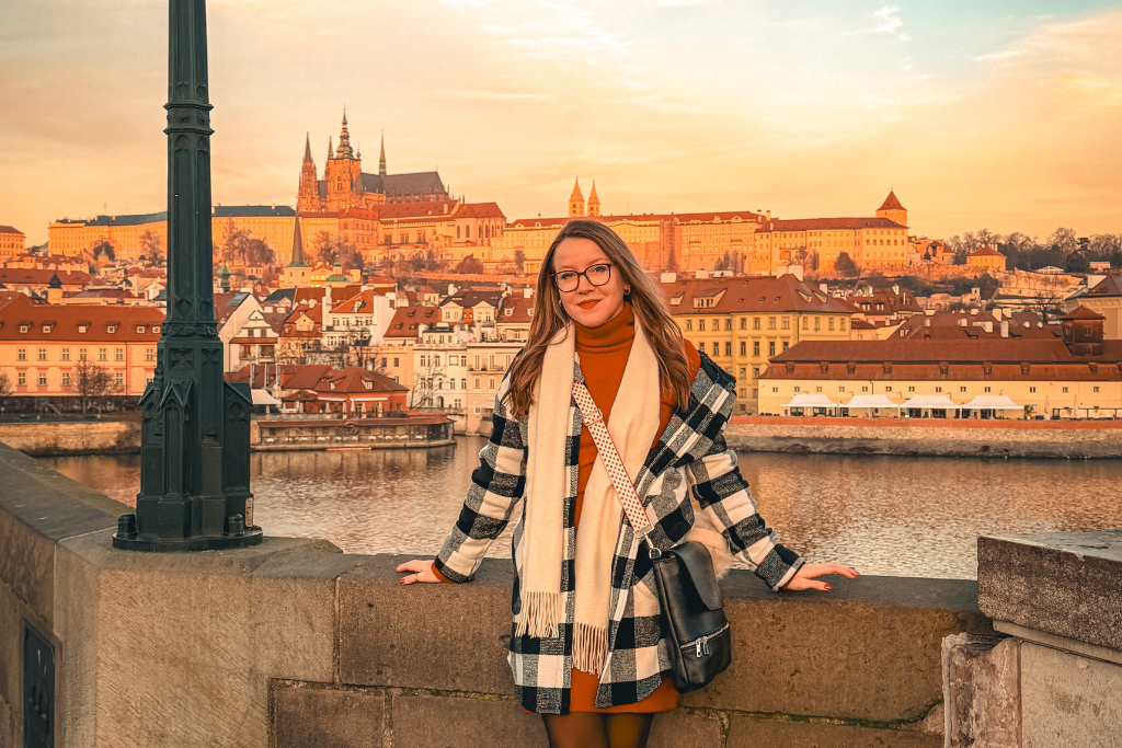Kate posing along the Vltava River with Prague Castle and St. Vitus Cathedral glowing in the background at sunset.