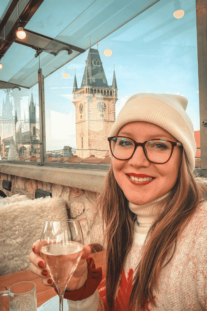 Kate enjoying a glass of wine at a rooftop terrace in Prague, with the Old Town Hall tower and Church of Our Lady before Týn in the background.