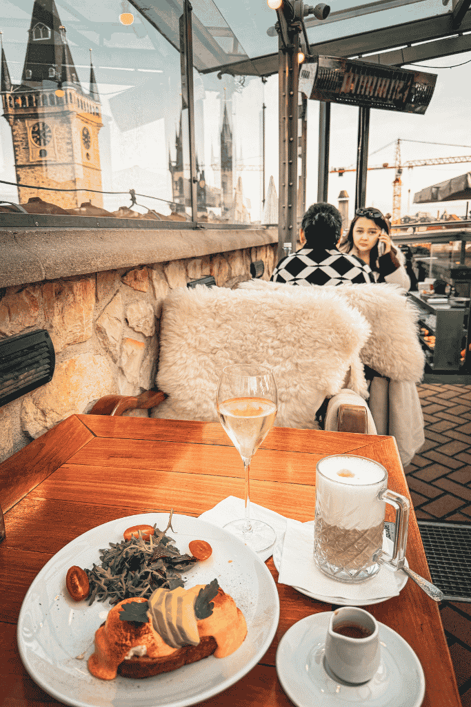 Breakfast on a rooftop terrace in Prague with coffee and wine, overlooking the Old Town Hall tower and historic city skyline.