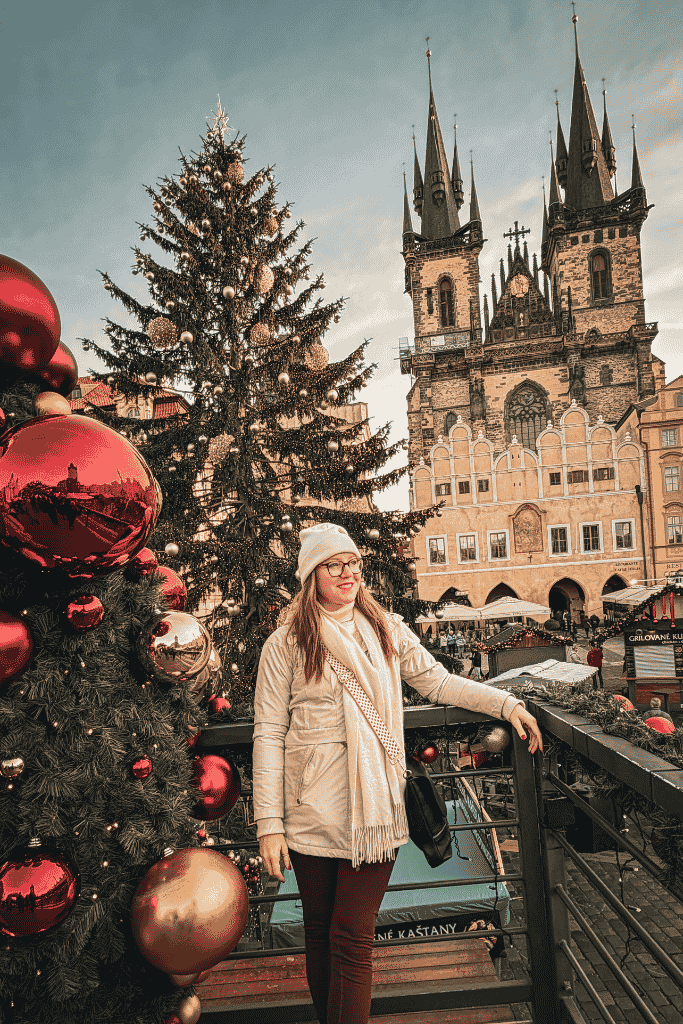 Kate posing beside a decorated Christmas tree overlooking Prague’s Old Town Square, with the Church of Our Lady before Týn in the background.