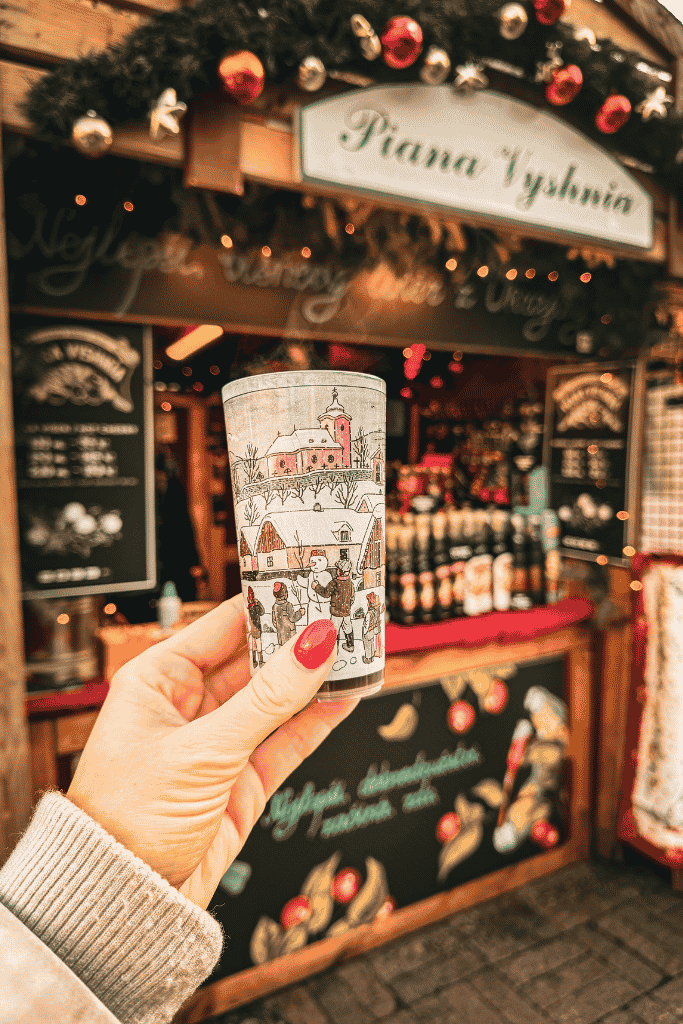 Hand holding a festive cup of hot mulled wine in front of a decorated Christmas market stall in Prague.