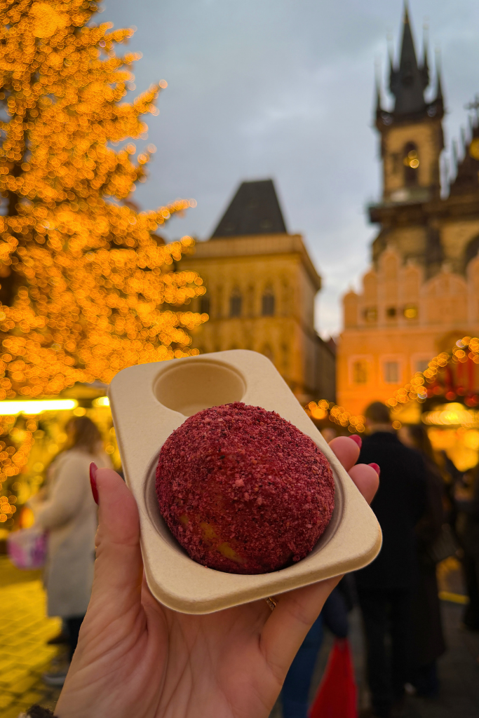 Sampling a sweet treat beneath the twinkling lights of Prague’s Christmas market, with the Gothic spires of Old Town Square glowing in the background.