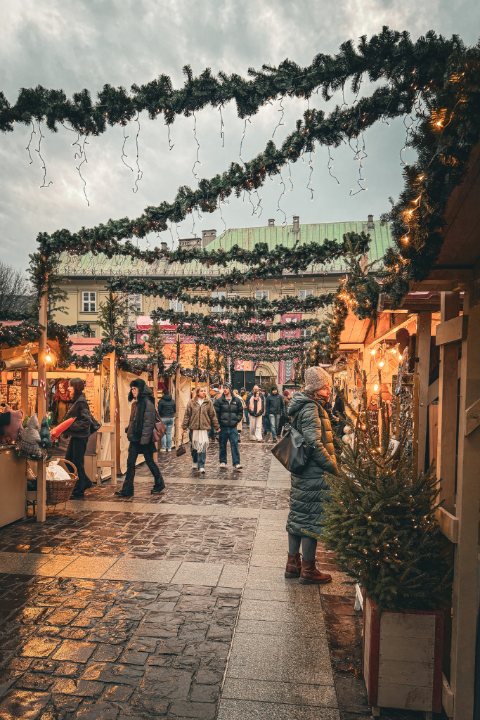 A cozy Christmas market in Krakow with festive garlands, glowing wooden stalls, and visitors strolling along the cobblestone street.