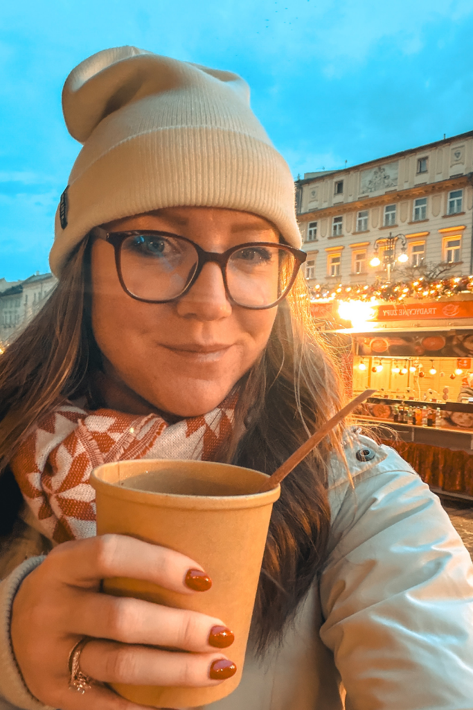 Kate wearing a white knit beanie, glasses, cream winter coat, and red-and-white scarf while sipping a hot drink at the Krakow Christmas Market.