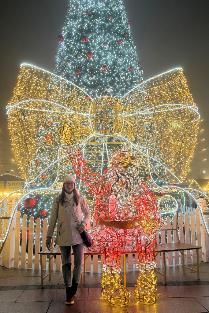 Kate posing beside a glowing Santa display and giant Christmas tree at Krakow’s train station Christmas Market.