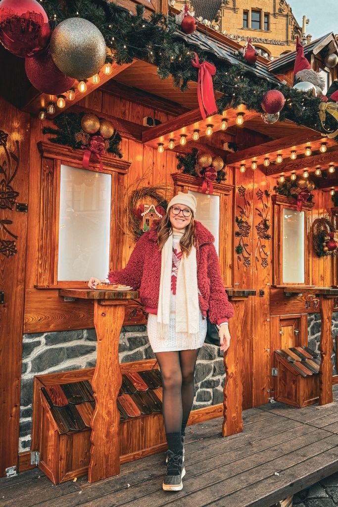 Kate eating at the Wroclaw Christmas Market beside a festive wooden stall decorated with lights and ornaments.