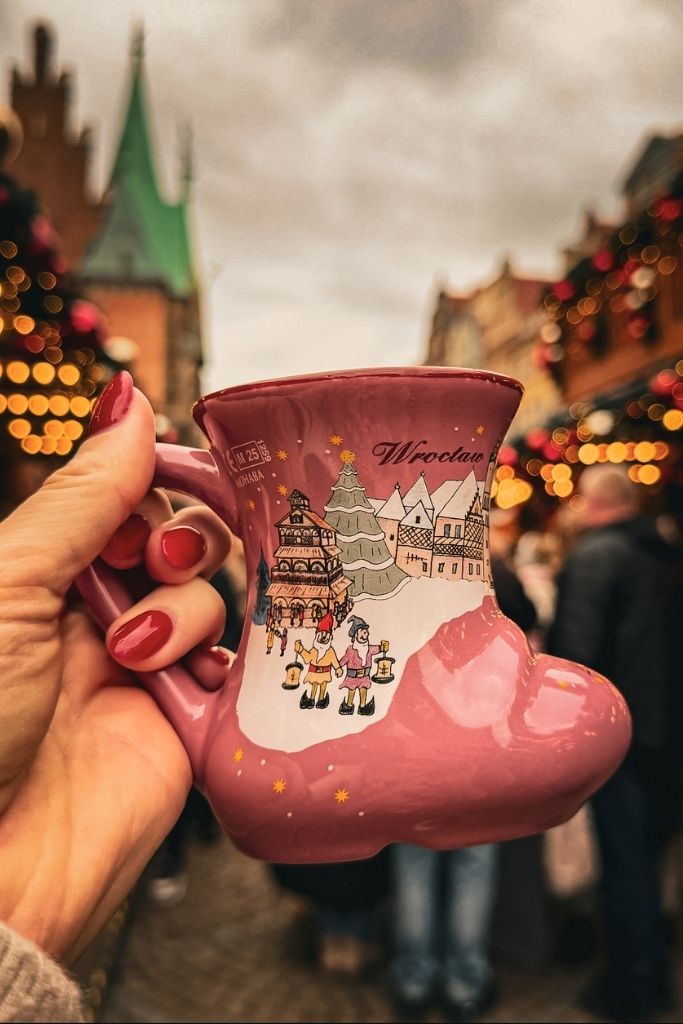 Pink boot-shaped collectible mug from the Wroclaw Christmas Market with festive stalls glowing in the background.