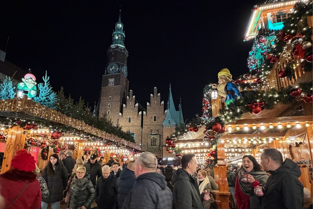 Wroclaw Christmas Market at night with glowing wooden stalls, festive decorations, crowds, and the illuminated Old Town Hall.
