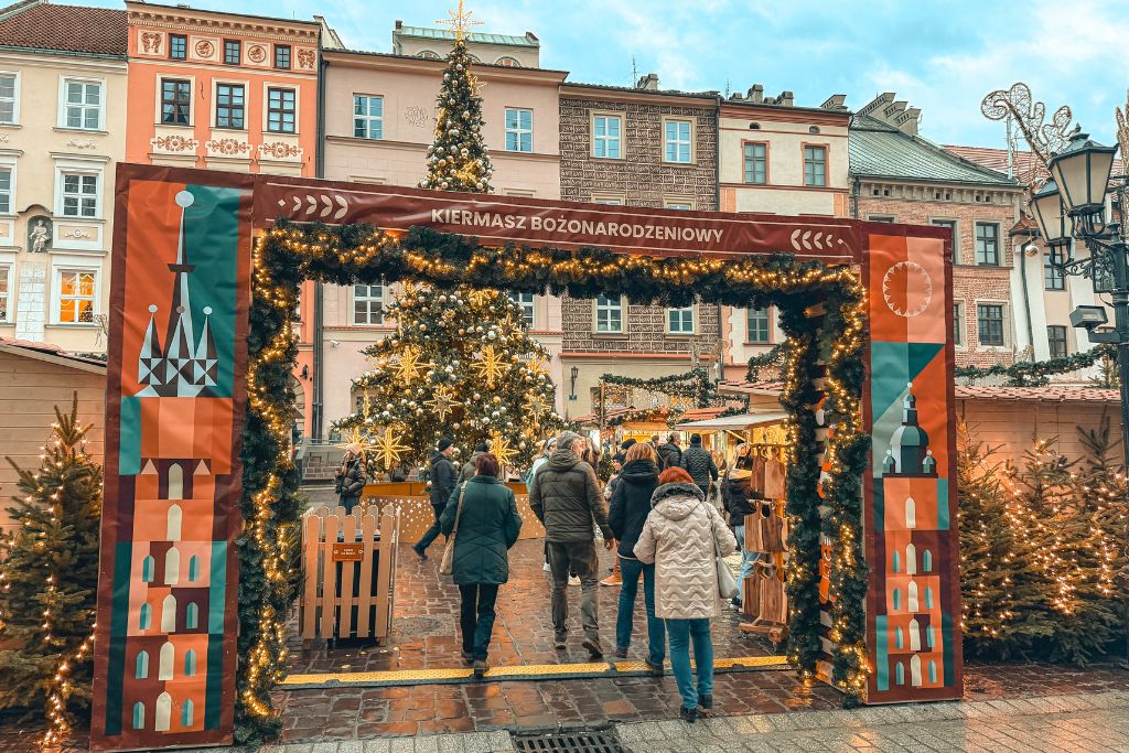 Entrance to the Little Market Square Christmas Market in Krakow with festive lights, Christmas trees, and wooden stalls.