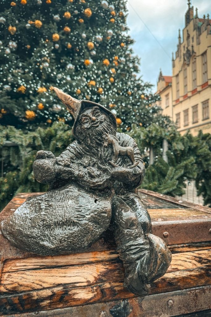 Wroclaw dwarf statue holding a sack with a decorated Christmas tree glowing in the background.