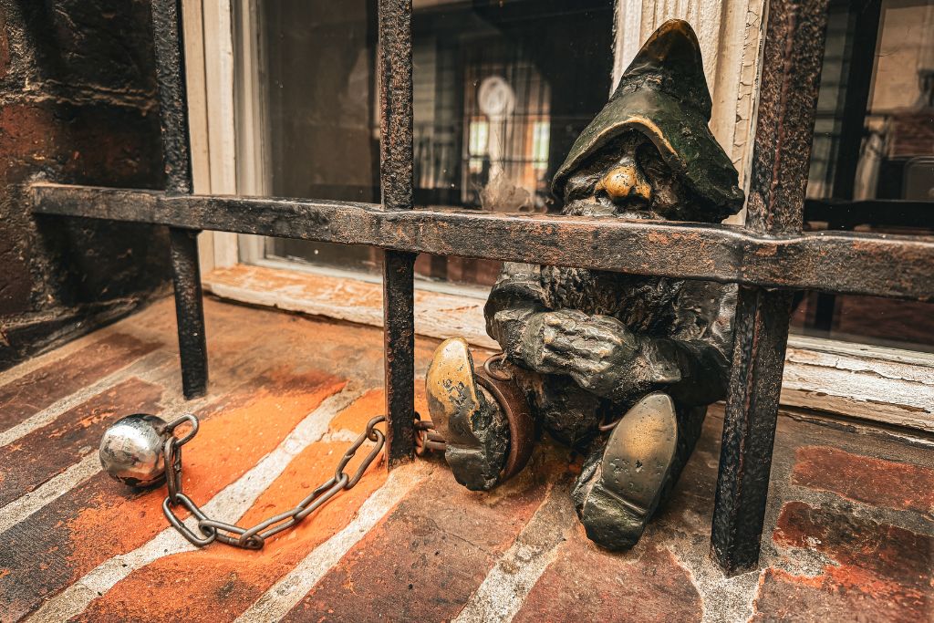 Wroclaw dwarf statue sitting behind metal bars with a ball and chain on a brick windowsill.