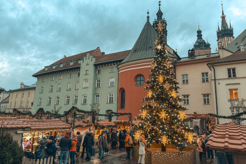 Little Market Square Christmas Market in Krakow at sunset, with a decorated Christmas tree, festive wooden stalls, and historic buildings in the background.